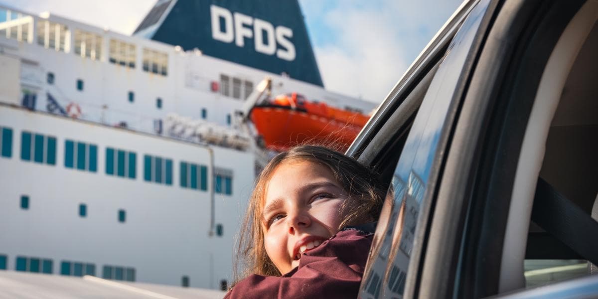 Excited little girl leaning out a car's window, with in the background a DFDS passenger vessel