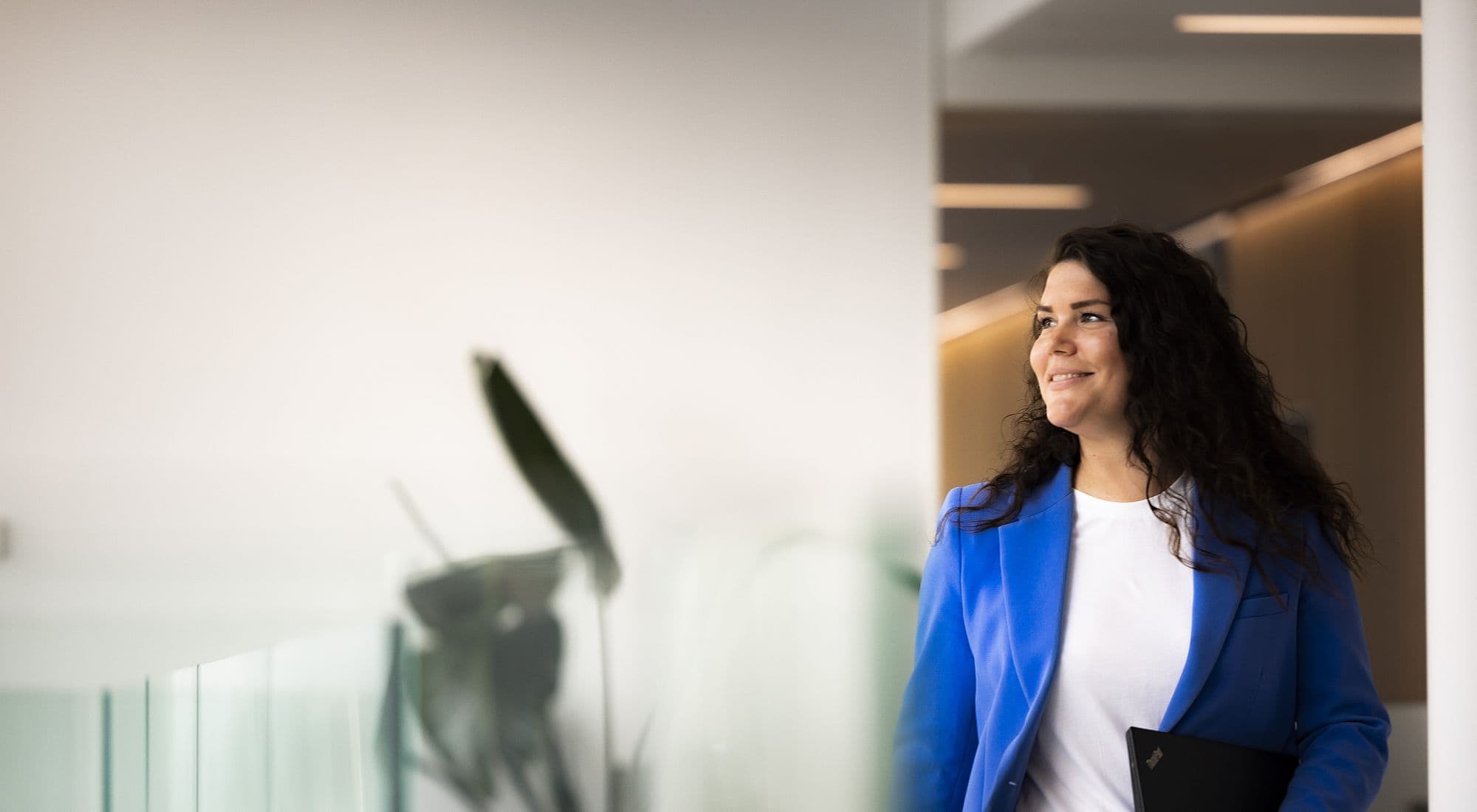 Hallway in new DFDS headquarter, woman in white shirt and blue jacket, walking with laptop in left hand, smiling