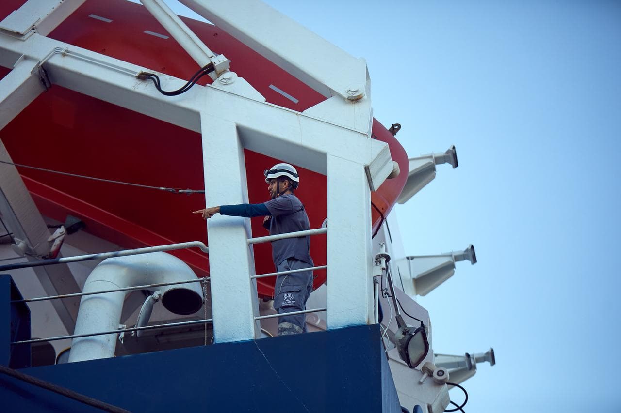 A seafarer with a white safety helmet on, standing on the deck of a DFDS vessel, pointing out a specific situation to other non-visible colleagues