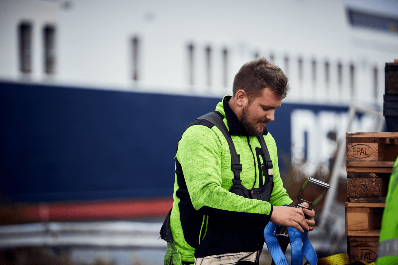 A terminal  with a yellow security jacket, working in the harbour with wooden pallets, within the background, a DFDS vessel