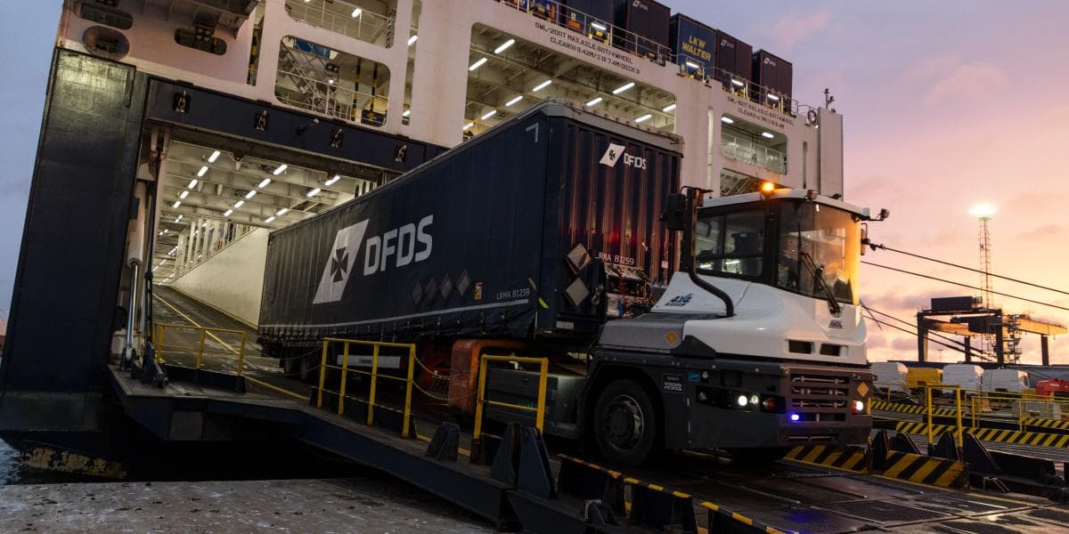 A white DFDS terminal tractor, with the lights turned on, pullying a darkblue DFDS trailer from a RoRo vessel in the harbour.