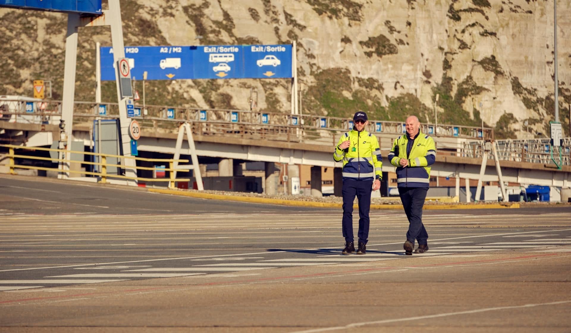 Two men wearing yellow security jackets walking in the harbour on the road.