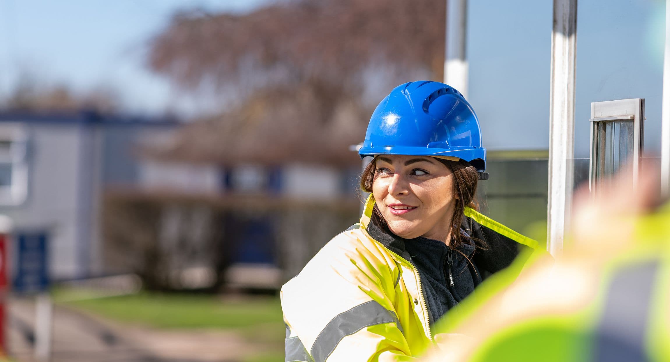 A woman standing outside in the sun, wearing a blue helmet and a yellow safety jacket, smiling and looking over her right shoulder backwards