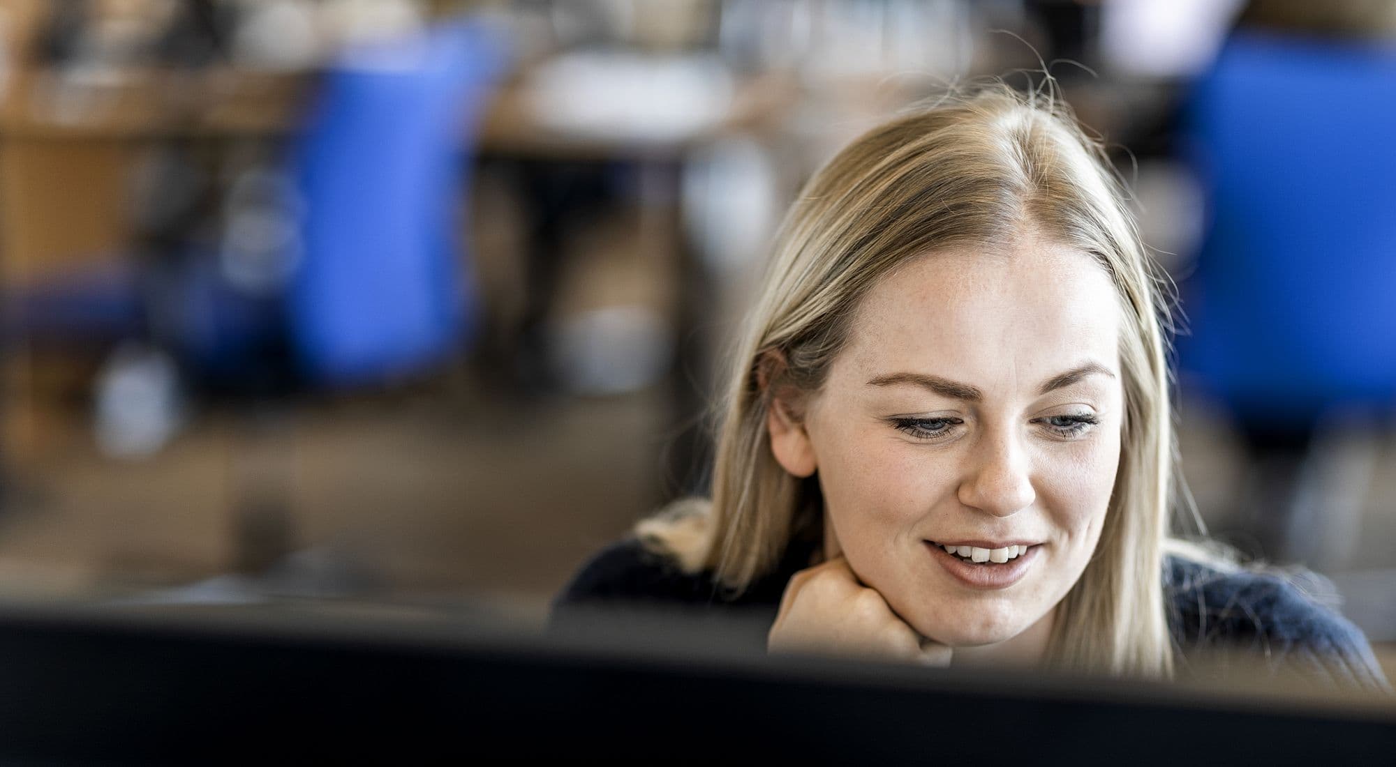 Woman sitting behind a desk in an office, looking at PC monitor, background blurred