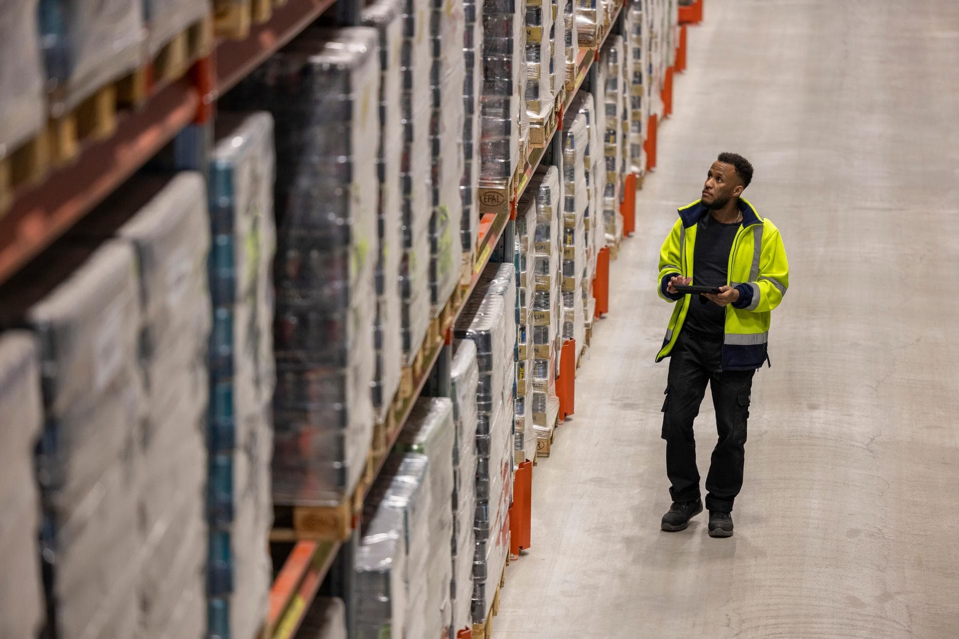 Man in a yellow jacket, standing in the warehouse before loaded storage rack, with a tablet in hand