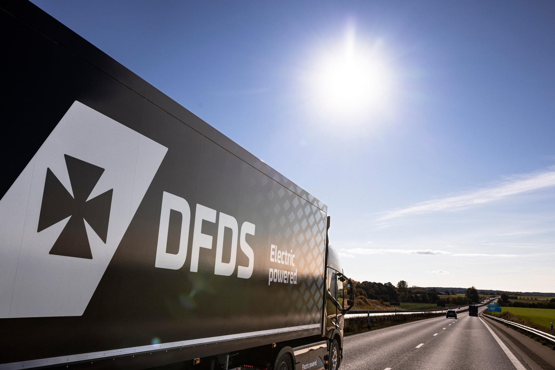 Electric DFDS truck, in blue colours, driving on the left lane of the highway, with sun high in the sky