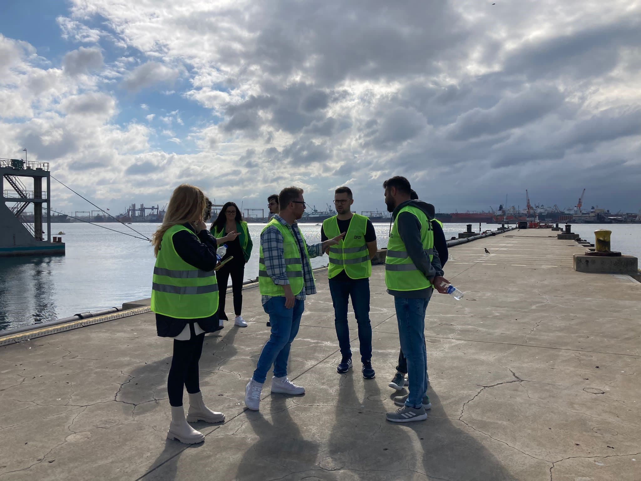 A group of men and women, standing on a harbour dock, wearing safety vests, standing together, discussing